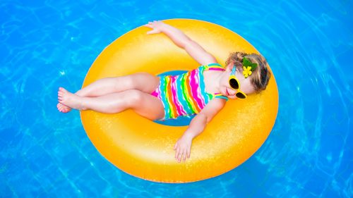 Cute funny little toddler girl in a colorful swimming suit and sun glasses relaxing on an inflatable toy ring floating in a pool having fun during summer vacation in a tropical resort